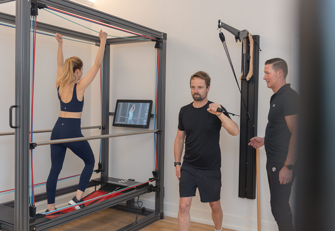 Zwei Personen trainieren an einem Gerät, der Trainer steht daneben. Zwei Personen trainieren an einem Gerät, der Trainer steht daneben.